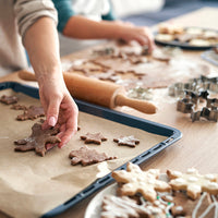 A close-up lifestyle shot of a person's hand placing a gingerbread cookie on a baking tray lined with parchment paper, with a wooden rolling pin and metal cookie cutters on a wooden table.