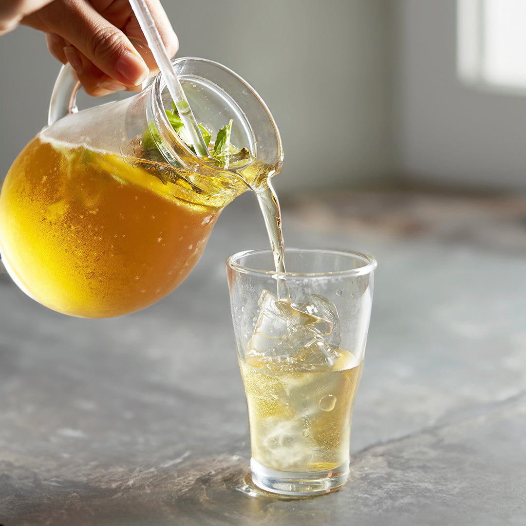 A hand pours a clear glass pitcher filled with a golden iced beverage into a clear glass tumbler with ice, set on a gray stone surface.