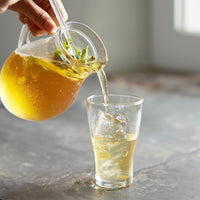 A hand pours a clear glass pitcher filled with a golden iced beverage into a clear glass tumbler with ice, set on a gray stone surface.