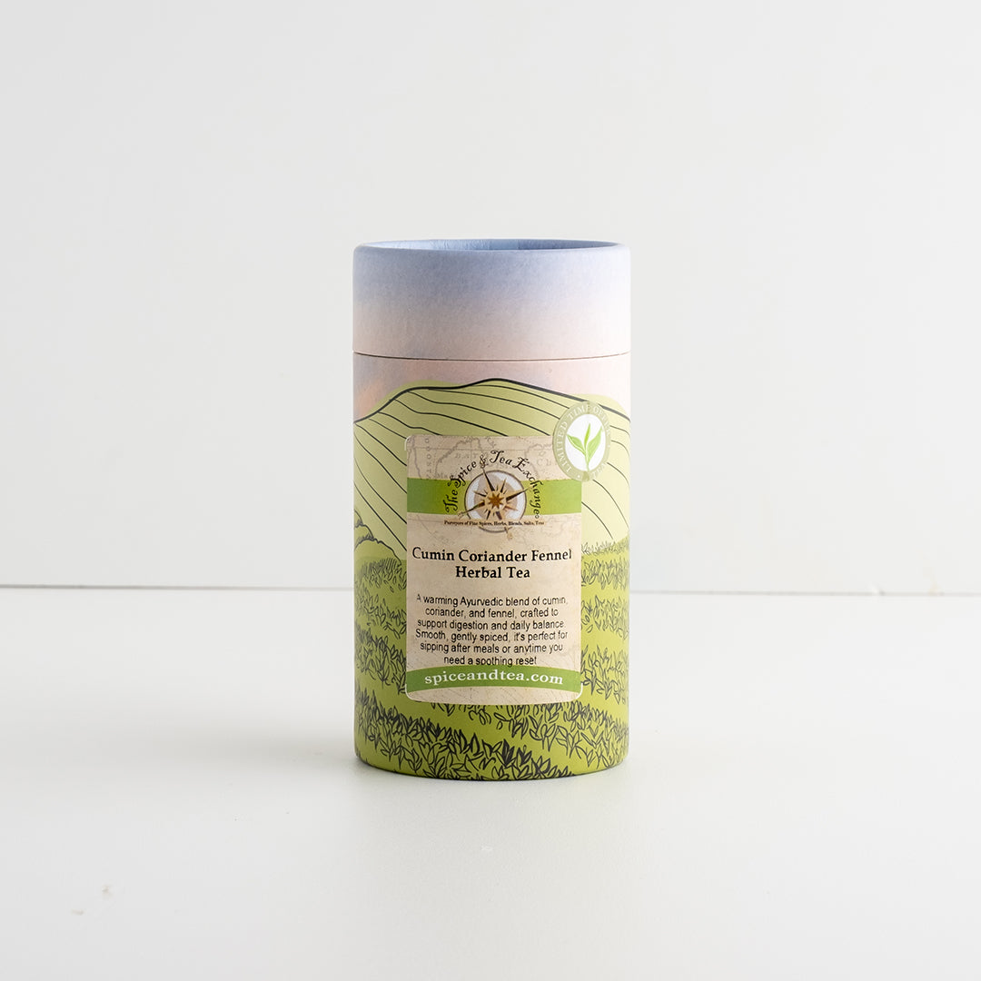 A cylindrical paperboard tea canister with a printed landscape design in green, beige, and soft blue, labeled 'Cumin Coriander Fennel Herbal Tea'