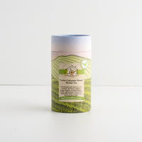 A cylindrical paperboard tea canister with a printed landscape design in green, beige, and soft blue, labeled 'Cumin Coriander Fennel Herbal Tea'