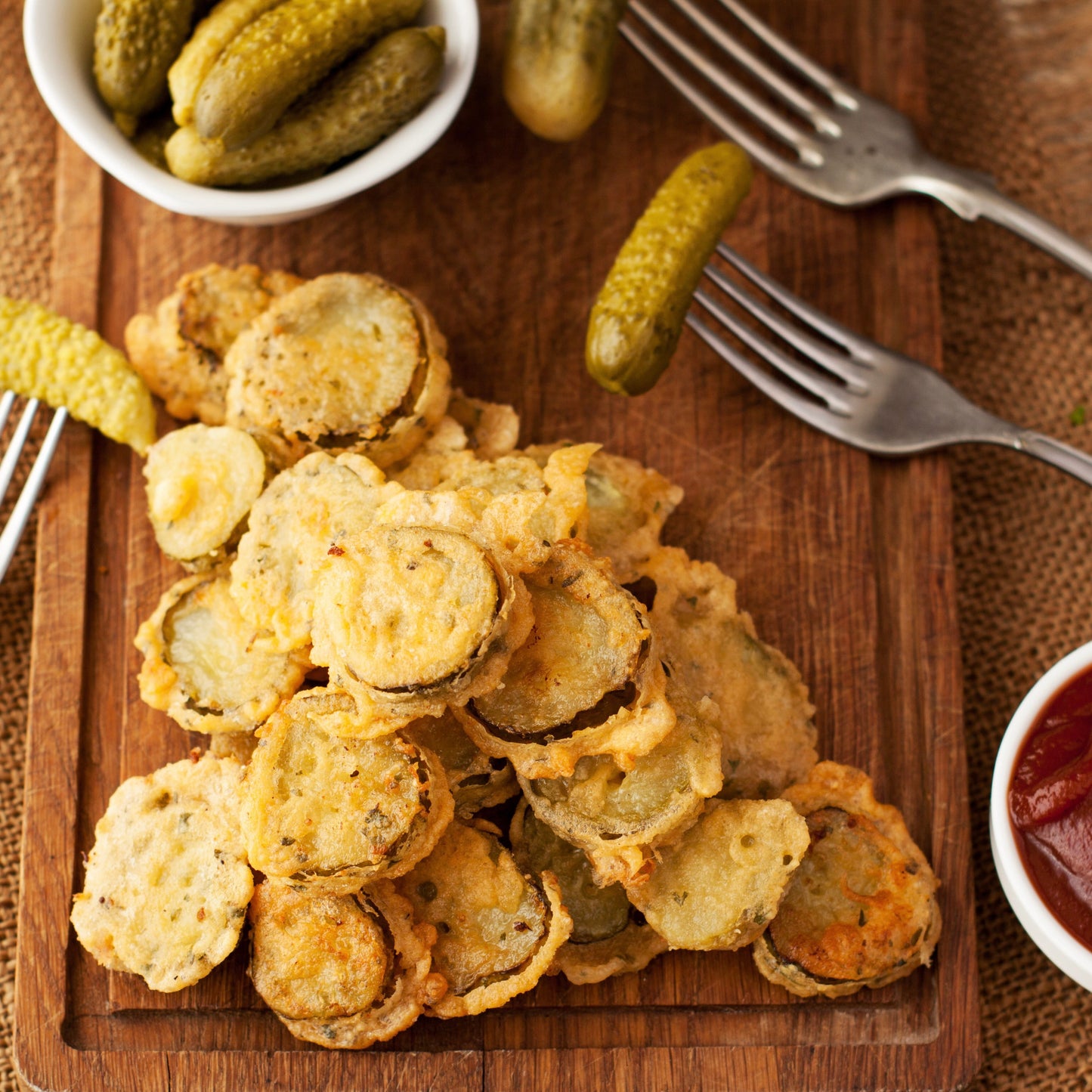 Golden-brown fried pickle slices piled on a wooden board, with whole pickles, forks, and small bowls of sauce around them on a rustic table.