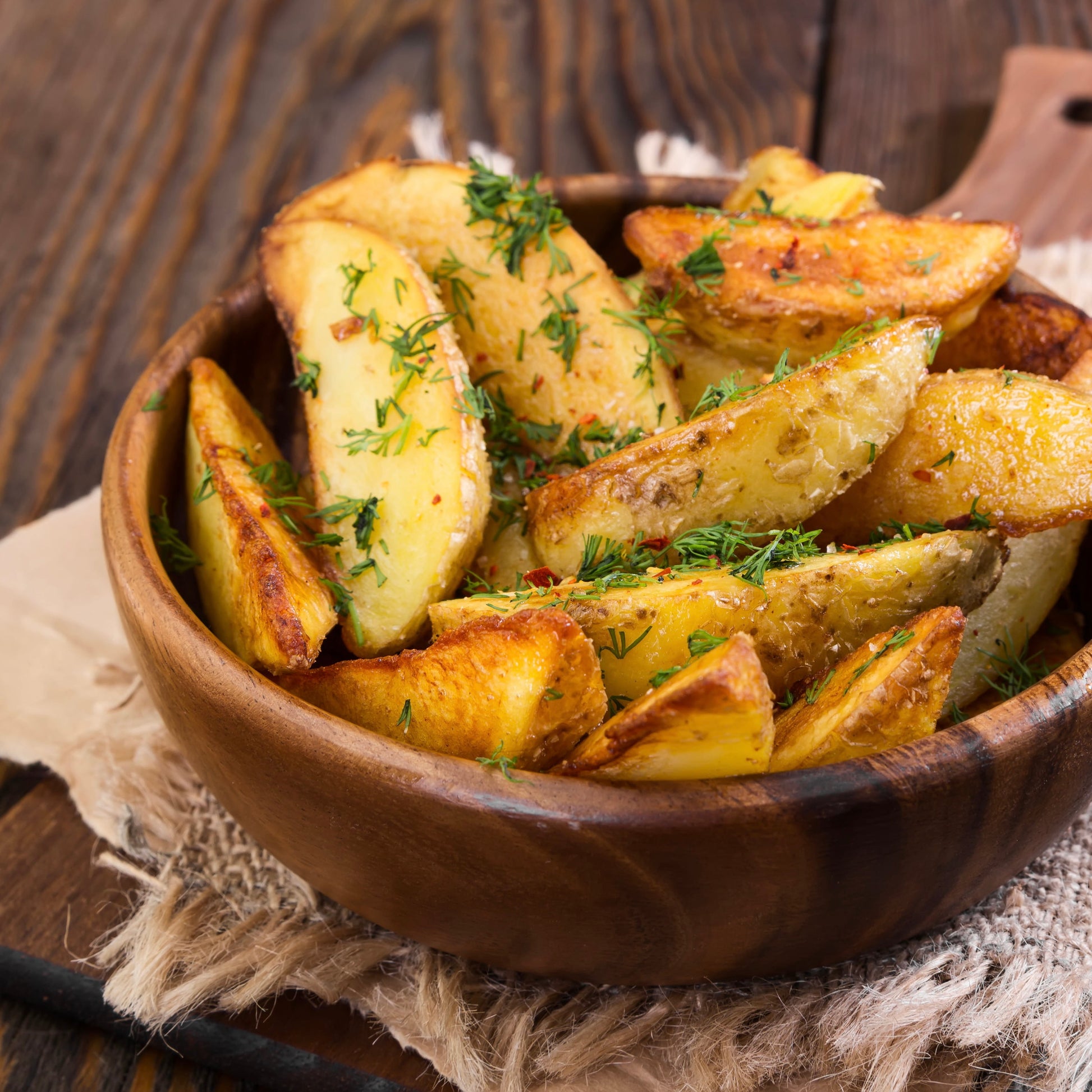 Roasted potato wedges in a wooden bowl, garnished with chopped green herbs and spices, set on a rustic wooden table with a burlap cloth underneath.
