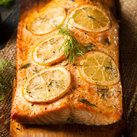 A close-up of a baked salmon fillet topped with lemon slices and dill and seasoning on a wooden serving board over burlap.