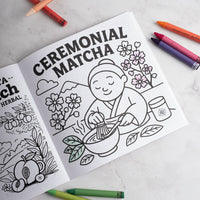 Coloring book open to a page with 'Ceremonial Matcha' text and illustrations, surrounded by crayons on a light surface.