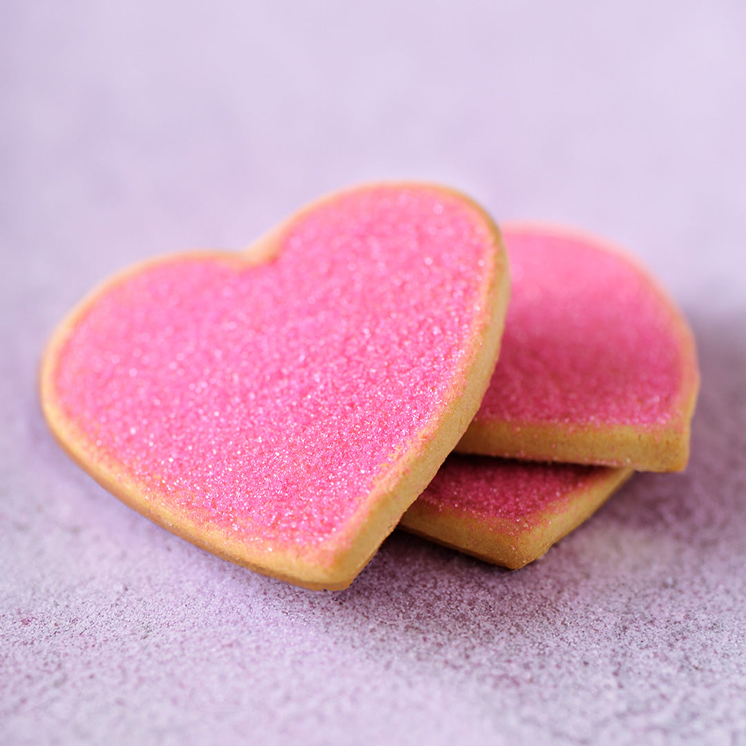 Three heart-shaped sugar cookies dusted with pink sugar, stacked on a soft lavender background.