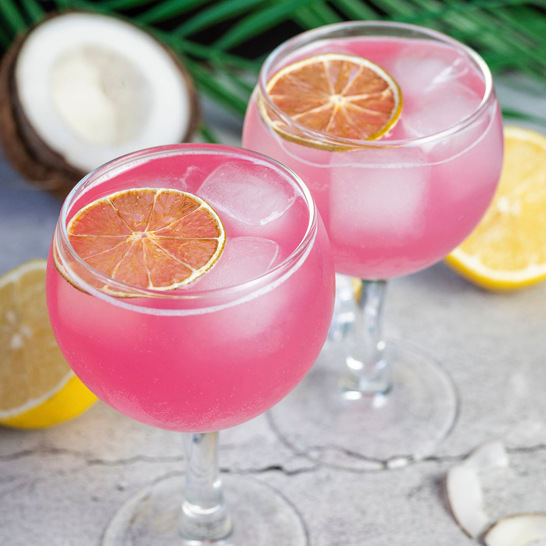 Two pink cocktails in clear stemmed glasses with ice, each topped with a citrus slice, styled on a stone surface with lemons, coconut, and green leaves in the background.
