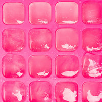 A close-up of a bright pink silicone or plastic ice cube tray with multiple square compartments, some containing translucent pink ice cubes.