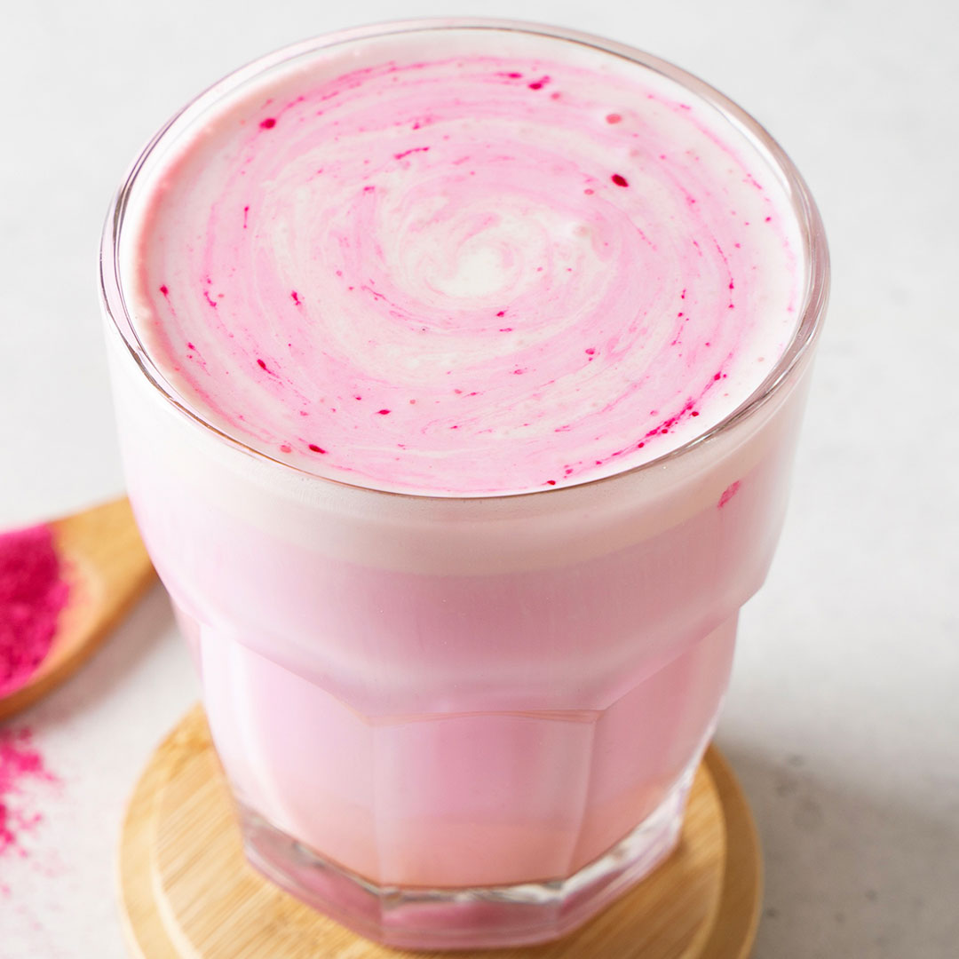 A pink and white swirled drink in a clear faceted glass, resting on a small wooden coaster with a wooden spoon of bright pink sugar nearby on a light surface.