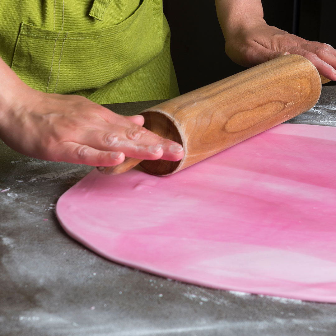 A person wearing a green apron uses a wooden rolling pin to roll out a sheet of pink dough on a floured gray surface.