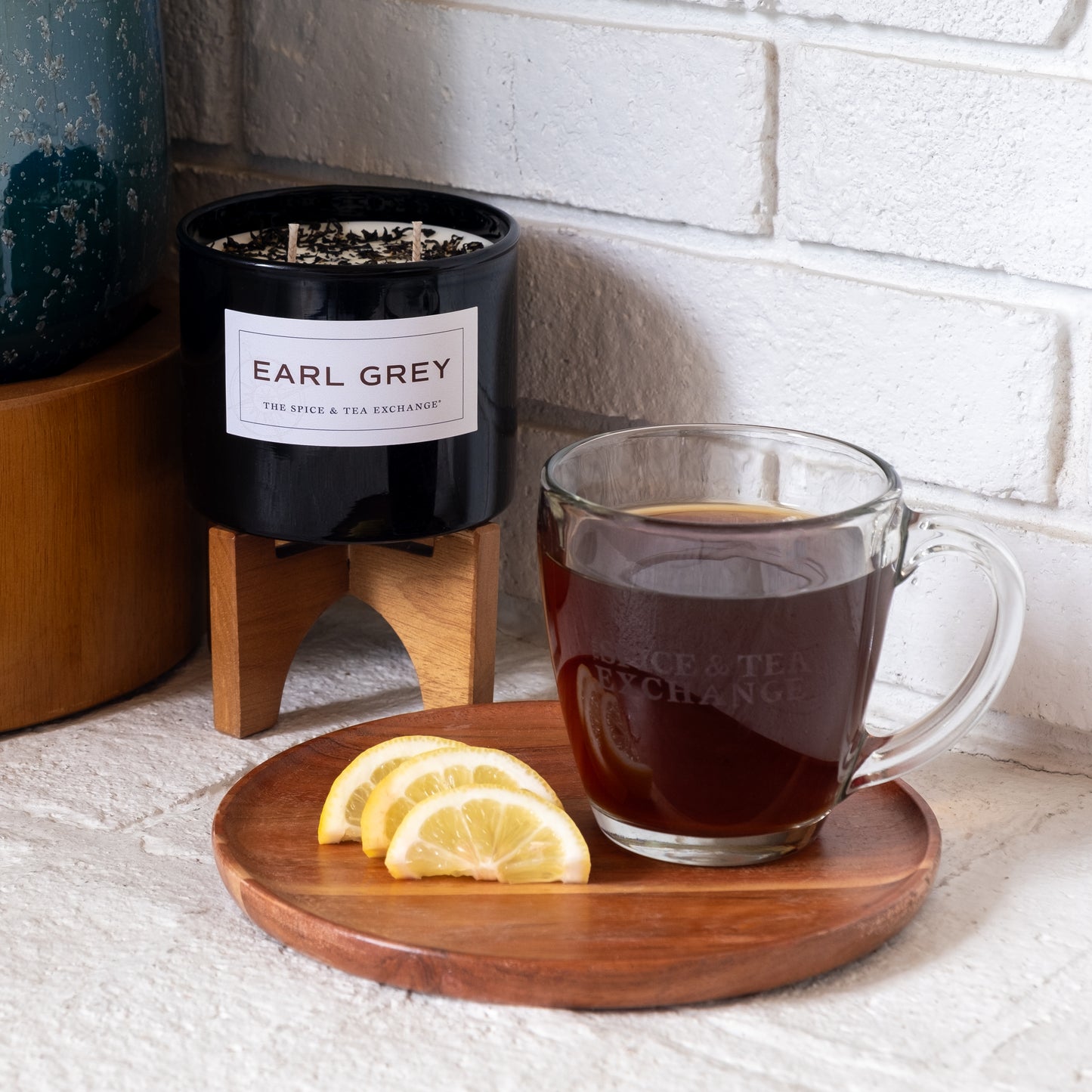 A glass mug of Earl Grey Black Tea sits on a wooden plate with three lemon slices, beside an Earl Grey Candle in a black container on a wooden stand, all set against a white brick wall scented with its aromatic fragrance.