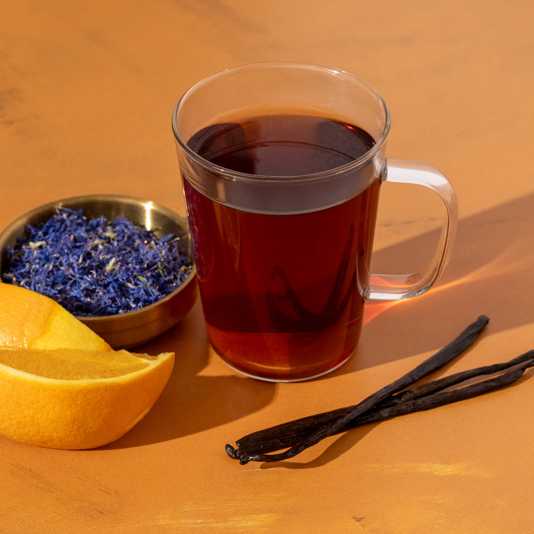 Earl Grey Creme Tea in glass mug next to vanilla beans, orange slices, and blue flower petals