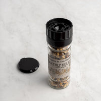 A glass jar of Earthly Delight Spice Blend with a black grinder top sits on light marble, ready to spice up meats, stews, and soups. The jar’s black cap rests nearby.
