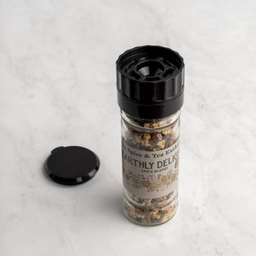 A glass jar of Earthly Delight Spice Blend with a black grinder top sits on light marble, ready to spice up meats, stews, and soups. The jar’s black cap rests nearby.