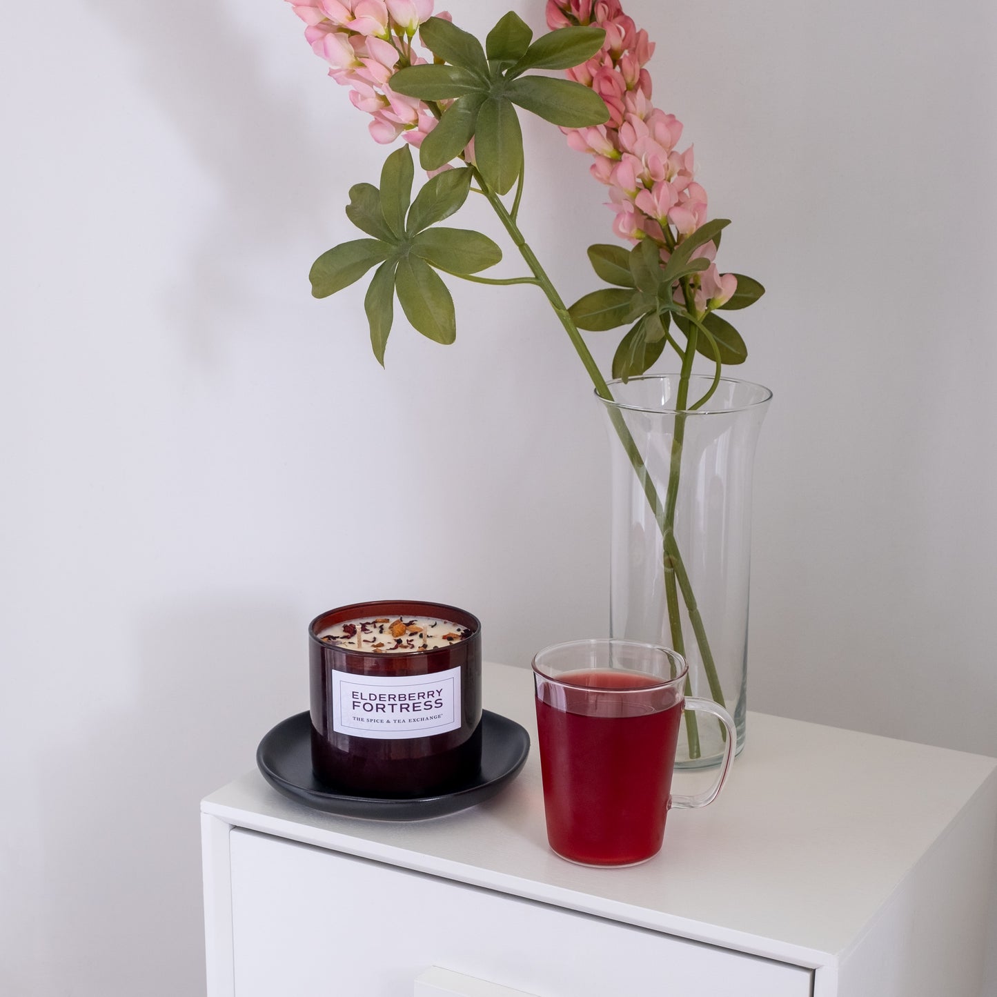 An Elderberry Fortress Candle in dark red soy sits on a white cabinet with a matching red drink in a glass and a tall vase of pink flowers, all set against a plain wall.