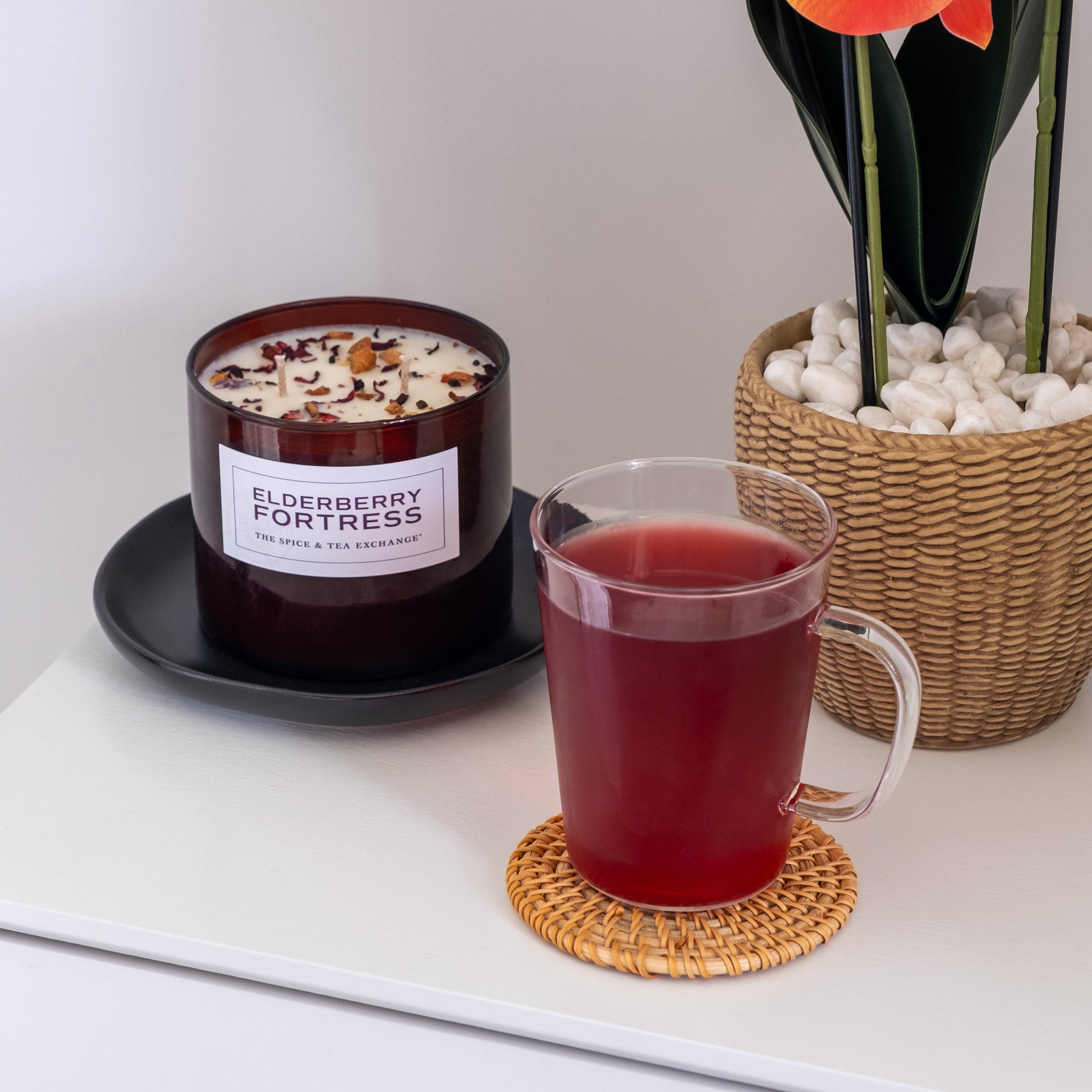 A glass mug of deep red elderberry tea rests on a woven coaster next to an Elderberry Fortress Candle made from natural soy and essential oils, with a potted plant on a white tabletop.
