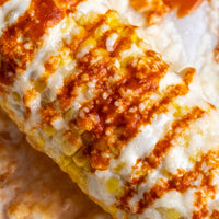 Close-up of corn topped with melted white cheese and topped with orange-red sauce, showing corn kernels and a crispy textured surface.