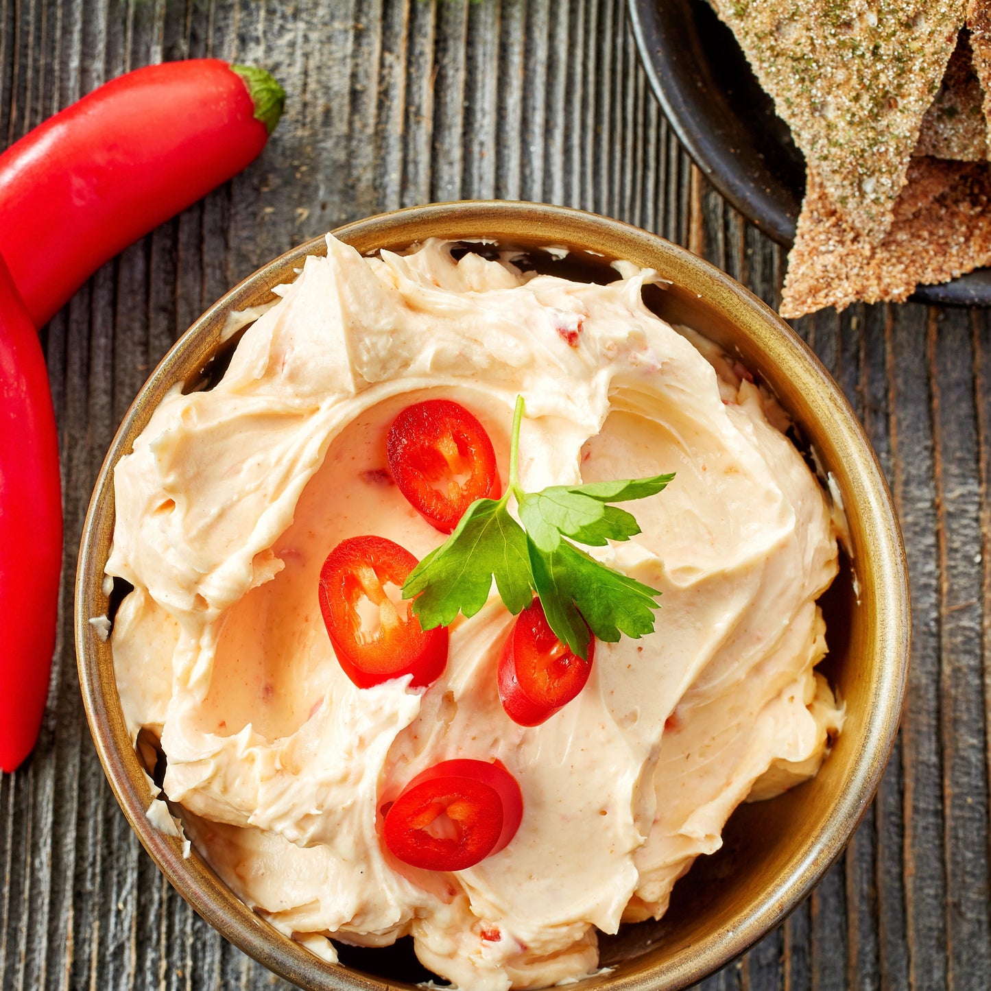 A top-down view of a creamy, light orange-beige dip or spread in a brown ceramic bowl, garnished with fresh red tomato slices and a green parsley sprig, set on a dark rustic wooden table with red chili peppers and crackers nearby.