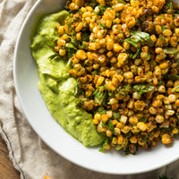 A close-up overhead shot of a white bowl filled with grilled corn kernels mixed with chopped herbs, set on a beige cloth over a wooden surface.