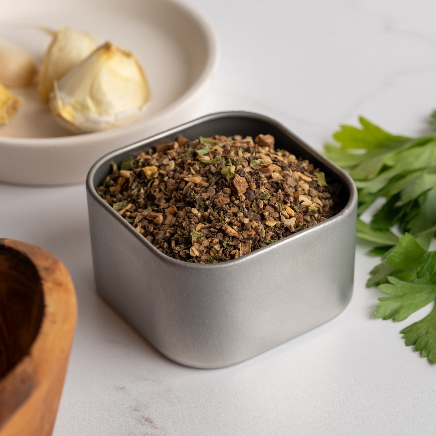 A small metal tin of Espresso Steak Rub sits on a white surface near fresh parsley, a garlic clove on a plate, and part of a wooden bowl, ready to add deep, rich flavor to your dishes.