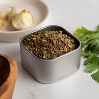 A small metal tin of Espresso Steak Rub sits on a white surface near fresh parsley, a garlic clove on a plate, and part of a wooden bowl, ready to add deep, rich flavor to your dishes.