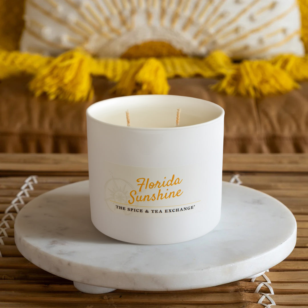 The Florida Sunshine Candle, a white soy candle on a round marble tray, fills the air with citrusy notes. A mustard yellow textured pillow and brown couch in the background add to the cozy atmosphere.
