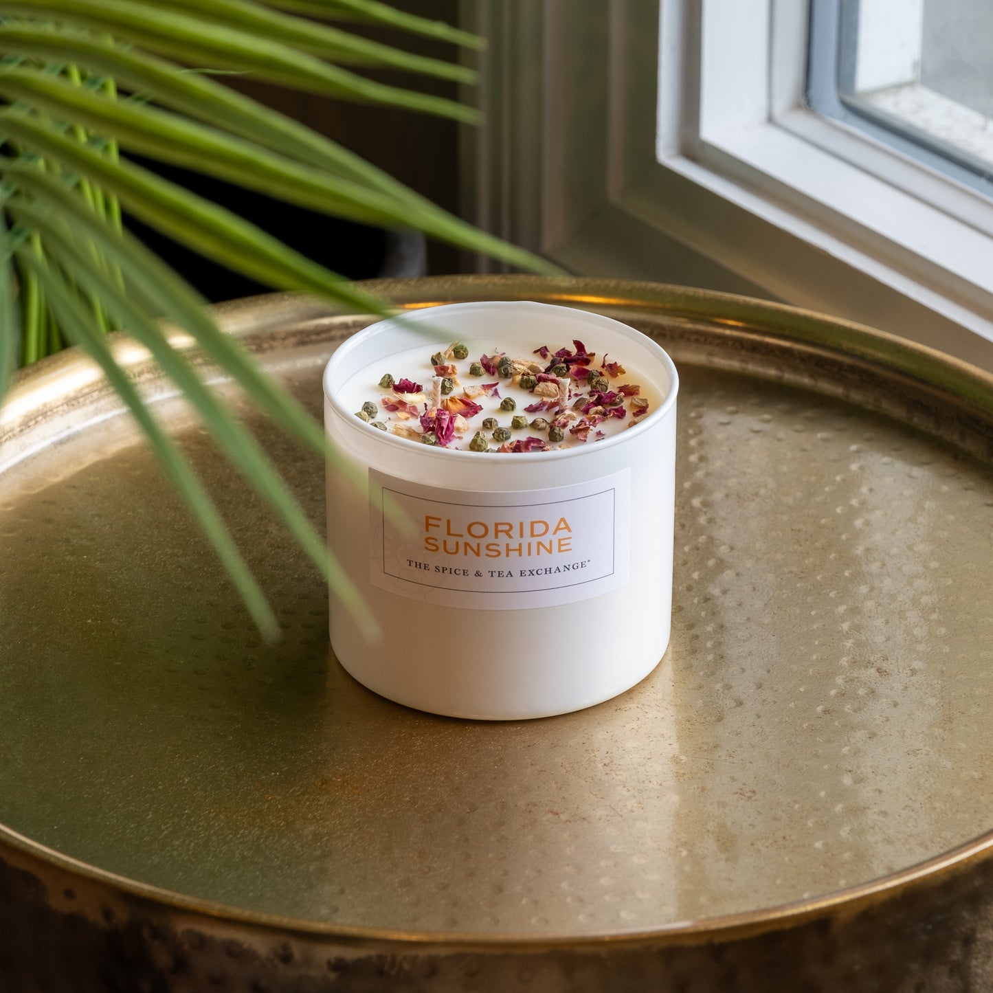The Florida Sunshine Candle, a white artisan soy candle, rests on a round gold tray near a window, topped with dried flowers and herbs. A green plant leaf appears in the foreground.