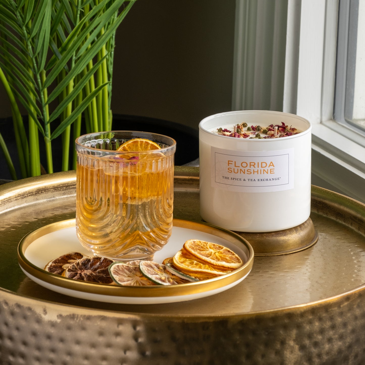 A glass of orange beverage with a dried citrus slice, assorted dried citrus slices, and a white Florida Sunshine Candle sit on a gold tray near a window and a green leafy plant.