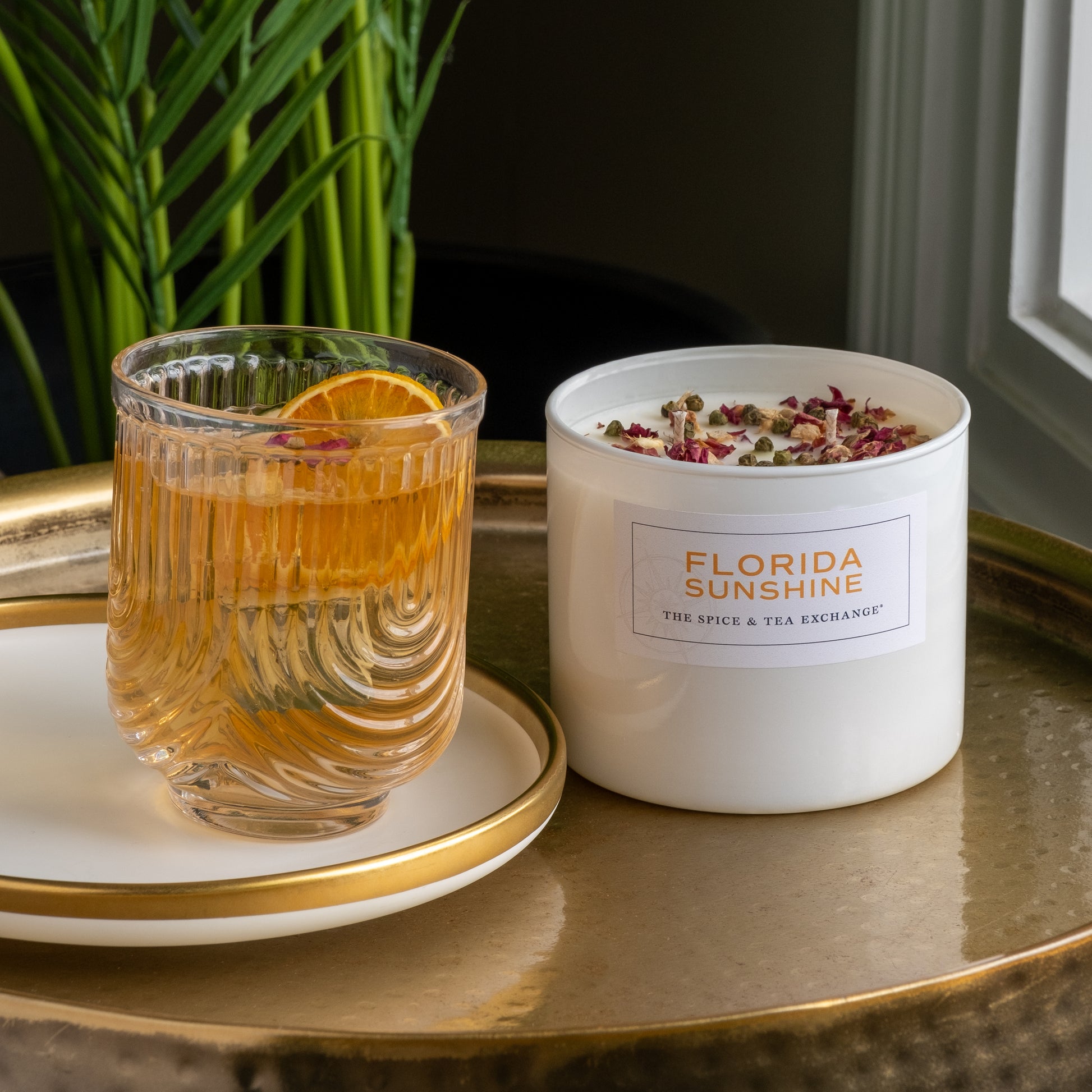 A glass of iced tea with an orange slice sits on a white and gold tray beside a Florida Sunshine Candle, an artisan soy candle. Green plants and a window appear in the background.