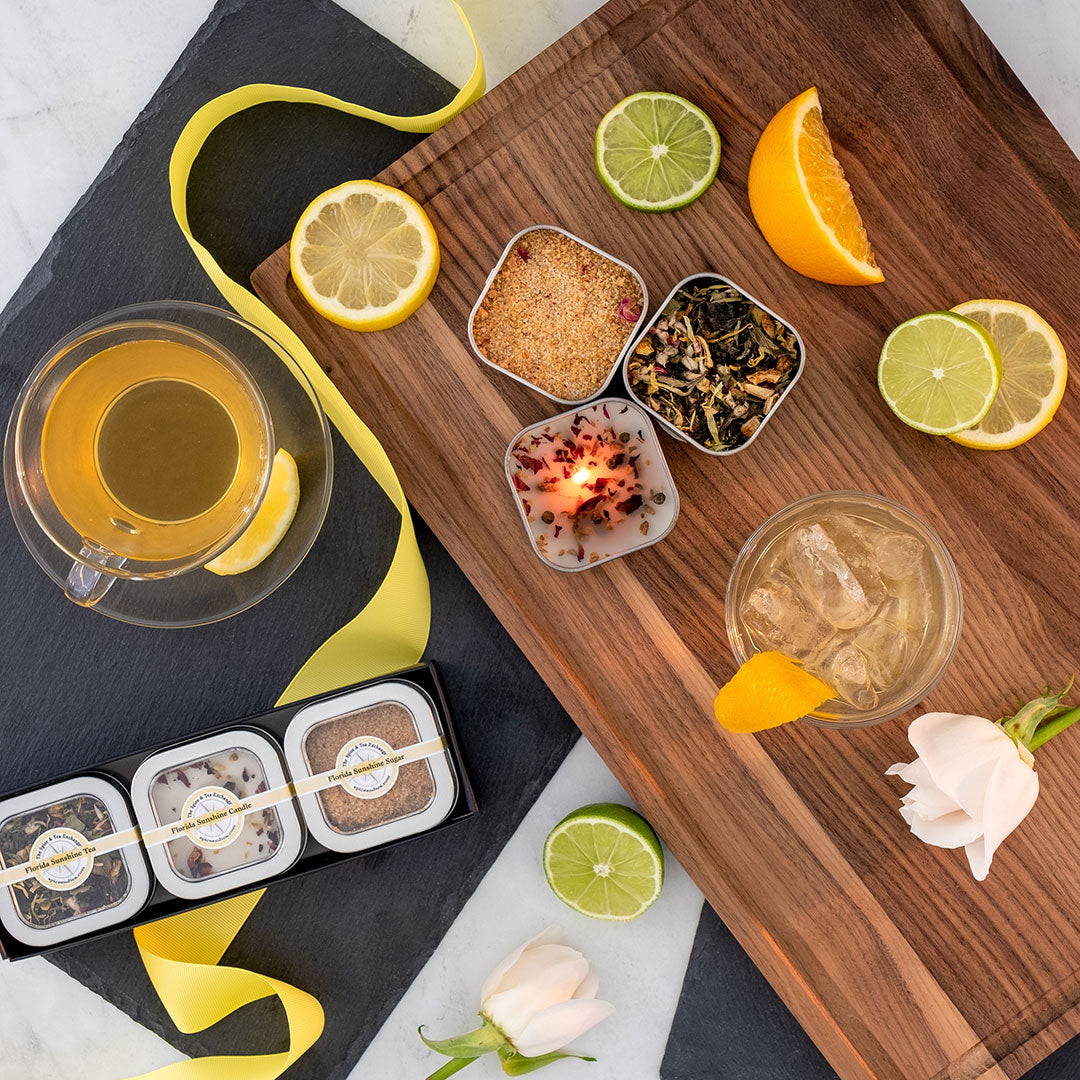 A wooden board with loose tea leaves, citrus slices, and brown sugar sits beside iced tea, hot tea, ribbon, roses, and the Florida Sunshine Tea Time Trio—an ideal marble-surface gift set for any tea lover.