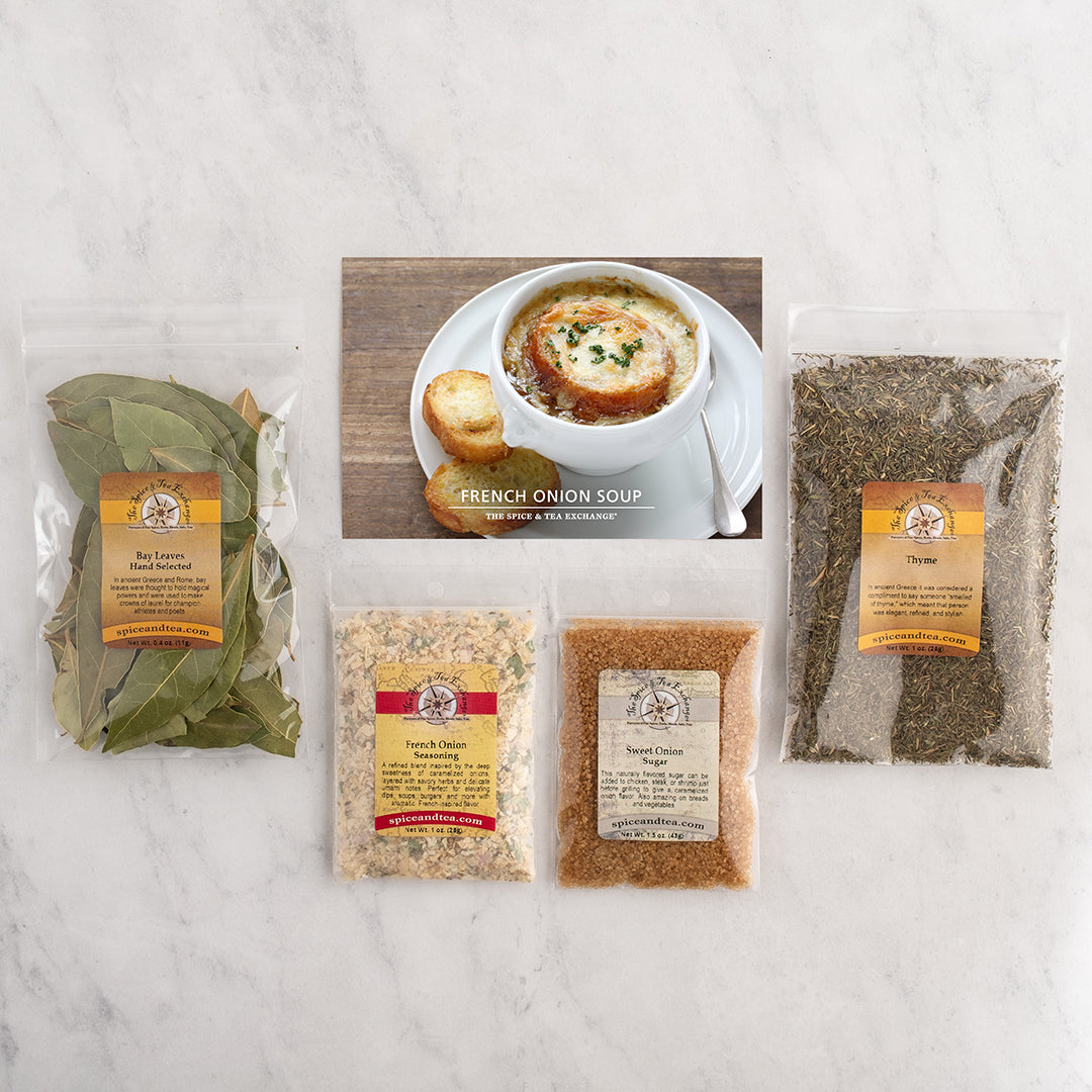 A French Onion Soup kit laid out on a white marble surface, featuring a bag of bay leaves, a bag of thyme, two bags of seasoning blends, and a recipe card showing a French onion soup dish.