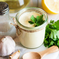 A small clear glass jar filled with creamy off-white sauce or dressing, tied with twine and topped with a parsley sprig, styled on a marble surface with garlic, parsley, a wooden spoon, and a salt shaker.