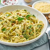 A close-up of a white speckled bowl filled with fettuccine pasta tossed in a creamy green pesto, topped with chopped parsley, walnuts, and parmesan cheese, with a fork and small bowls of ingredients in the background.