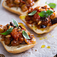 Three slices of toasted bread topped with a chunky tomato and eggplant sauce, garnished with fresh basil leaves, presented on crumpled parchment paper.