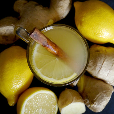 A glass mug filled with a pale yellow lemon ginger drink, topped with a lemon slice and a cinnamon stick, surrounded by fresh lemons and ginger roots on a dark background.