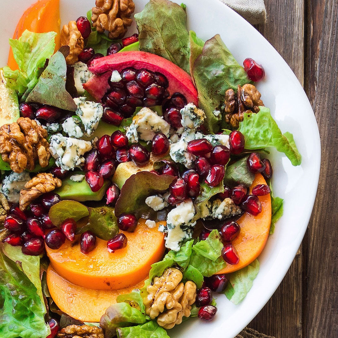 A close-up overhead shot of a fresh salad on a white plate, featuring mixed greens, sliced peaches, dark red pomegranate seeds, crumbled blue cheese, and walnuts on a rustic dark wood surface.