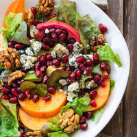 A close-up overhead shot of a fresh salad on a white plate, featuring mixed greens, sliced peaches, dark red pomegranate seeds, crumbled blue cheese, and walnuts on a rustic dark wood surface.