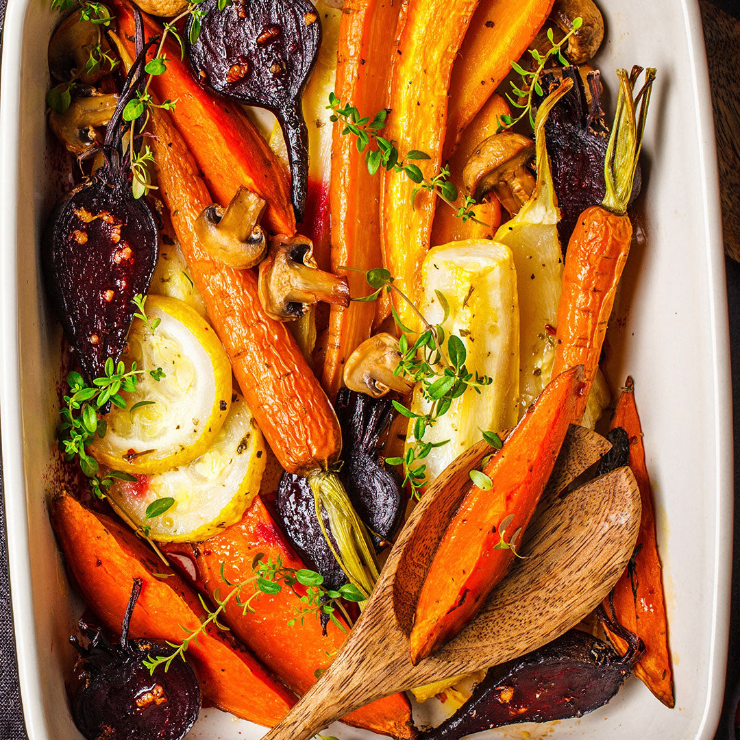 A close-up overhead shot of a white baking dish filled with roasted vegetables, including orange carrots, yellow onions, sliced lemons, brown mushrooms, and dark beets, garnished with fresh thyme and oil, with a wooden spoon resting on top.