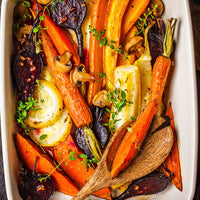 A close-up overhead shot of a white baking dish filled with roasted vegetables, including orange carrots, yellow onions, sliced lemons, brown mushrooms, and dark beets, garnished with fresh thyme and oil, with a wooden spoon resting on top.
