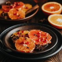 Cognac-infused orange slices garnished with pomegranate seeds and star anise, served on a dark ceramic plate on a wooden table, with more orange halves and a second plate blurred in the background.