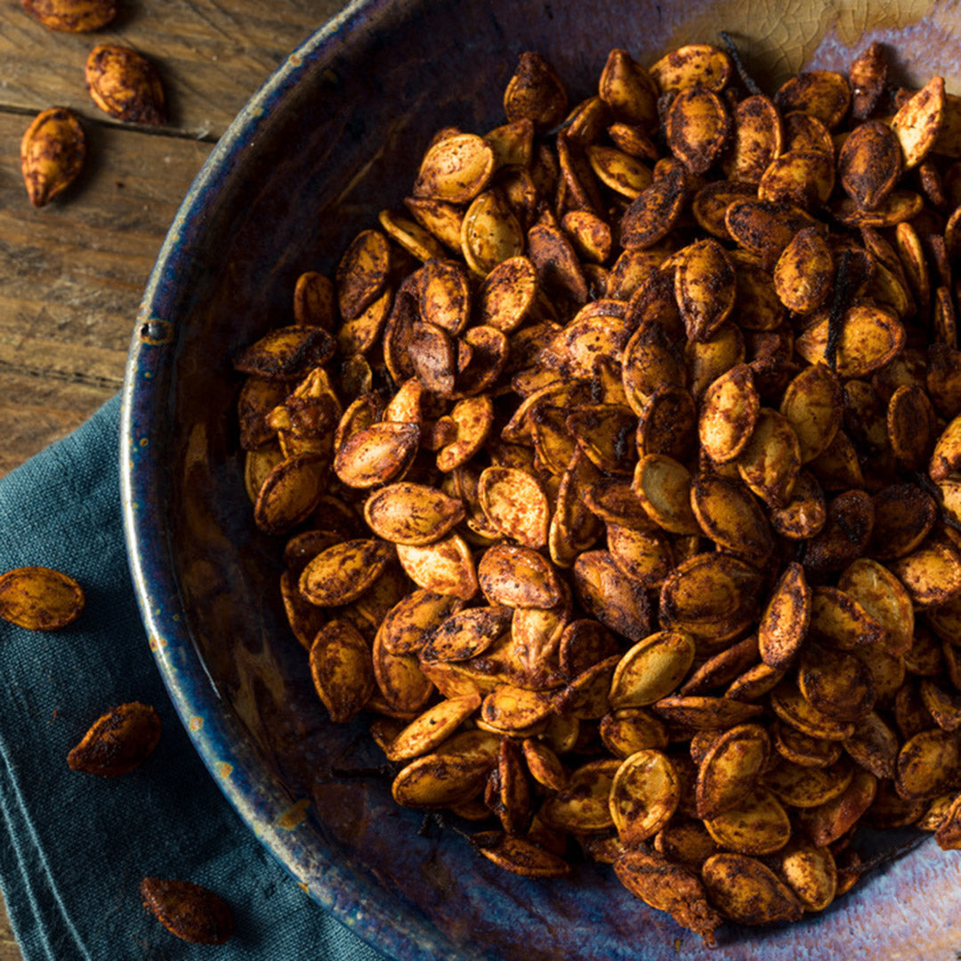 Crispy roasted pumpkin seeds in a rustic blue ceramic bowl, with a few scattered on a wooden surface and a dark blue cloth underneath.