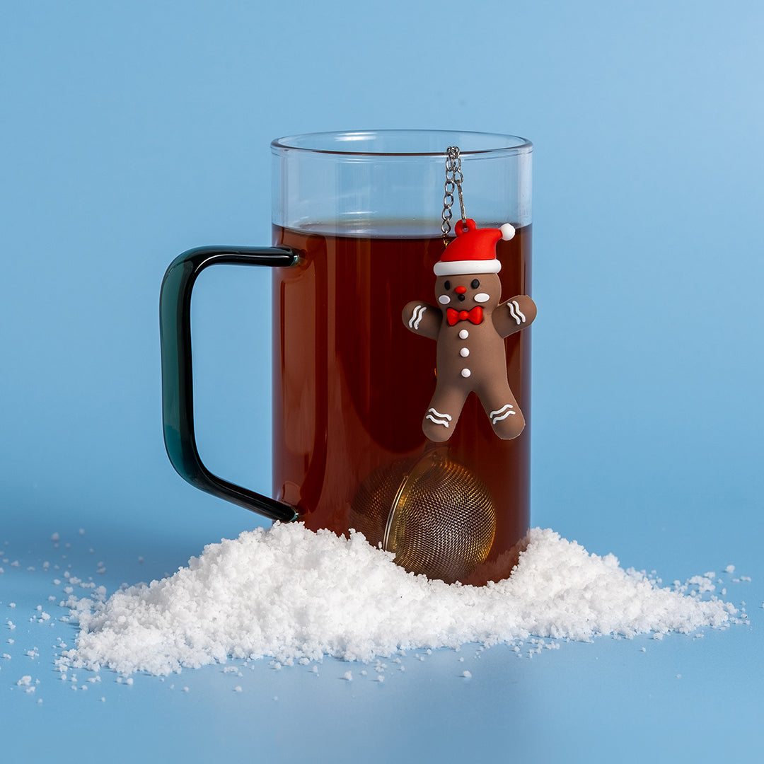 A clear glass mug filled with dark tea, featuring a gingerbread man-shaped tea infuser hanging from the rim, set on snow against a solid blue background.