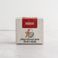 A square product box for The Spice & Tea Exchange Gingerbread Man Infuser, featuring a light gray marbled background, a red top label, and a gingerbread man illustration on a white marble surface.