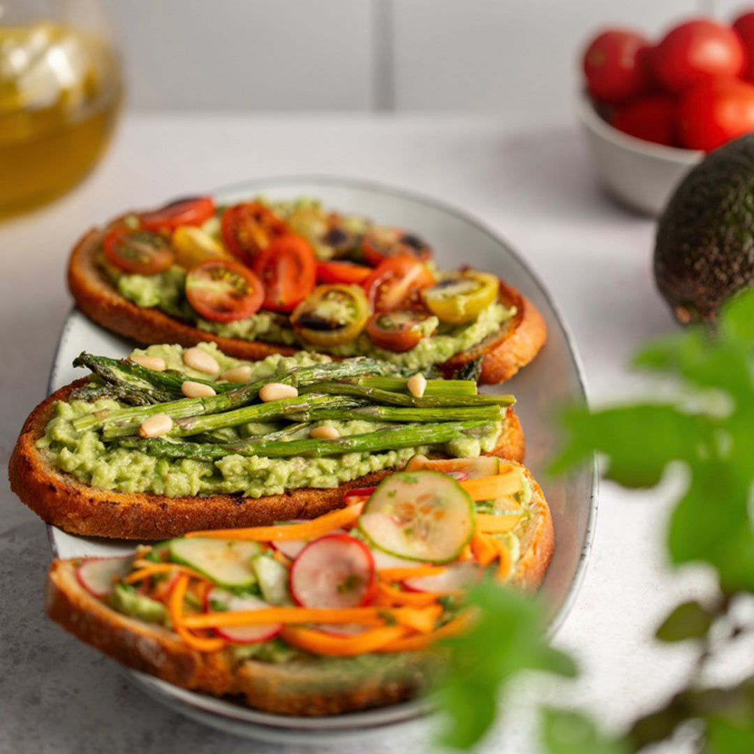 Three slices of bread with various toppings on a plate, including avocado, tomatoes, asparagus, and carrots.