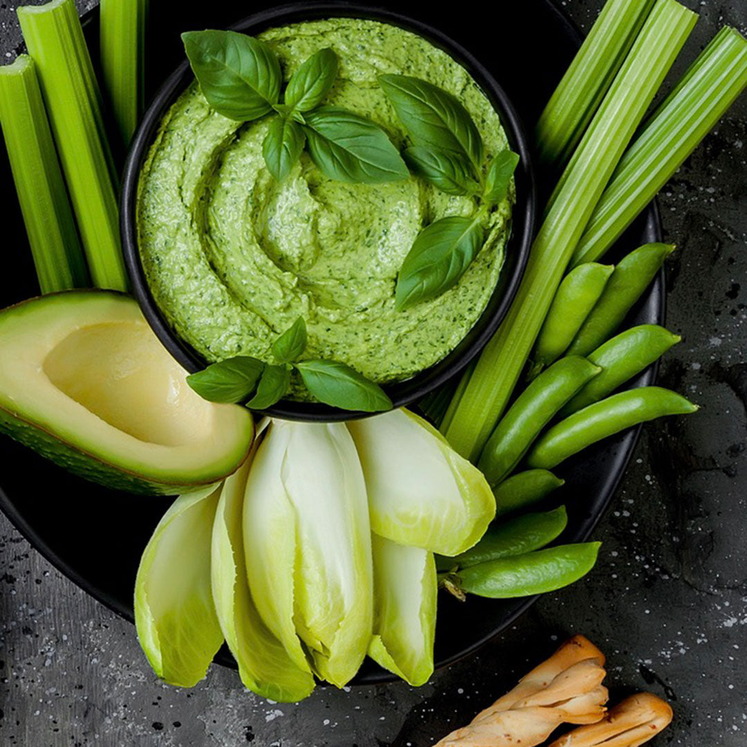 Green hummus with basil leaves surrounded by celery, avocado, and green beans on a dark surface.