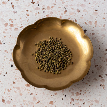A brass bowl with scalloped edges holds Green Peppercorn, valued for its herbal flavor, displayed on a terrazzo surface featuring pink and brown specks.