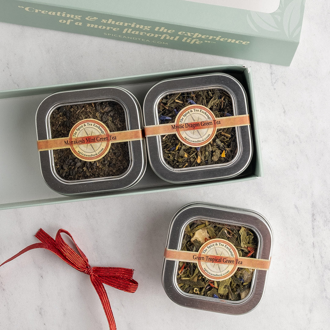 Three square metal tea tins displayed on a light marble surface with their open presentation box behind them. The tins are labeled as Herbal Tea with a tan ribbon. A red ribbon tied is sitting next to it.