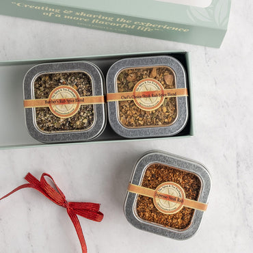 A gift set of three square metal spice tins displayed on a light marble surface, with one tin open to reveal two spice blends inside and next to a red ribbon. The tins have silver lids, tan paper bands, and round beige labels.