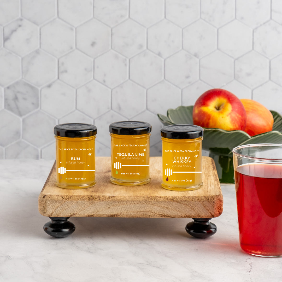 Three jars of honey on a wooden stand with a glass of red tea and apples in the background.