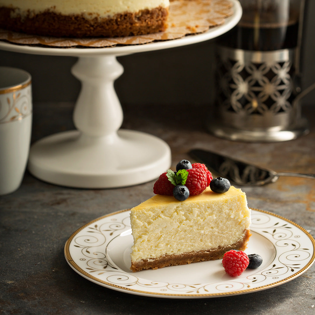 Slice of cheesecake with berries on a decorative plate, with a cake stand and coffee in the background.